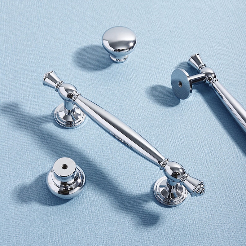 May include: A collection of polished chrome cabinet hardware, including two bar pulls, a cabinet knob, and a smaller knob. The hardware has a reflective surface and is arranged on a light blue surface.