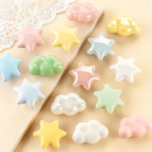 May include: A selection of pastel-coloured ceramic knobs in star and cloud shapes. The knobs are in shades of pink, yellow, blue, green, and white. These decorative knobs are ideal for adding a whimsical touch to furniture or decor.