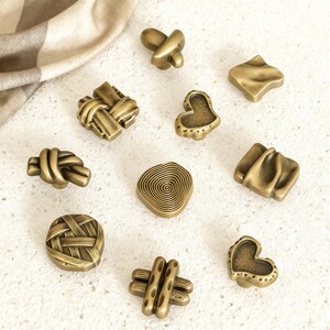 Puede incluir: Una colección de tiradores de armario de latón antiguo en varias formas, incluyendo un corazón, un nudo y un diseño tejido. Los tiradores están dispuestos sobre una superficie de color claro con una tela de tono neutro en el fondo.