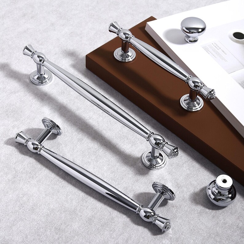 May include: Shiny chrome cabinet hardware, including two long bar pulls, a smaller bar pull, and two round knobs. The hardware is displayed on a brown and white surface, showcasing the reflective quality of the metal.
