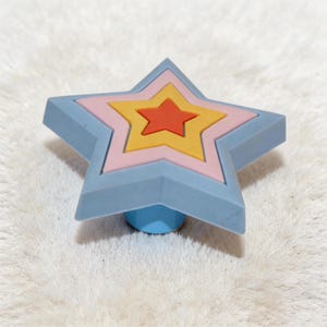 Op de afbeelding: Een meerlagig stervormig decoratief object. De buitenste laag is lichtblauw, gevolgd door roze, geel en een rode ster in het midden. Het object heeft een lichtblauwe basis en is waarschijnlijk een trekker of decoratieve knop.