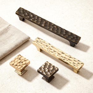 Op de afbeelding: Een assortiment kastbeslag, waaronder twee lange stangtrekkers en twee vierkante knoppen. Het beslag heeft een getextureerd, gehamerd ontwerp in brons- en goudtinten. Een neutraal gekleurde doek staat op de achtergrond.