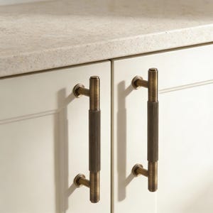 May include: Two antique brass cabinet pulls are shown, each with a vertical, ribbed centre section. They are mounted on a white cabinet door. The pulls are approximately 15 cm long, adding a touch of vintage charm to the cabinetry.
