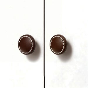 May include: Two brown leather cabinet knobs with white stitching. The knobs are round and have a scalloped edge. They are mounted on a white cabinet door.