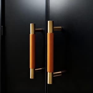 Pode incluir: Close-up de duas portas de armário pretas com ferragens a condizer. Cada porta apresenta uma pega dourada com uma pega cilíndrica revestida a couro laranja. As pegas são montadas horizontalmente, proporcionando uma estética elegante e moderna.
