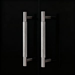 Könnte beinhalten: Zwei silberne Schrankgriffe mit strukturiertem, rautenförmigem Griff. Die Griffe sind zylindrisch und auf einer schwarzen Oberfläche montiert, was eine moderne und minimalistische Ästhetik schafft.