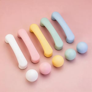 May include: A collection of colorful cabinet hardware, including six curved handles and three round knobs. The handles are in white, pink, yellow, green, and blue. The knobs are in white, pink, and yellow. The items are arranged on a pink surface.