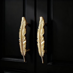 May include: Two gold-colored feather-shaped cabinet handles mounted on a black cabinet door. The handles are detailed with intricate feather vein patterns. The handles are made of metal and have a shiny finish.