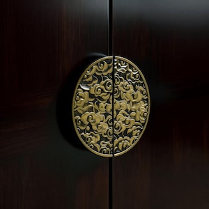 May include: A dark wood cabinet door with a decorative brass handle. The handle is circular with a cloud-like pattern. The brass handle is split in half, with each half on a separate door. The cabinet is dark brown.