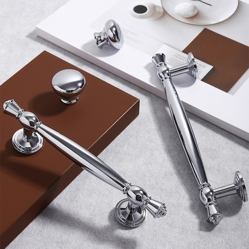 May include: A collection of polished chrome cabinet hardware, including two long bar pulls and two knobs. The pulls have a central, rounded grip and decorative end pieces. The knobs are a round, smooth design. The hardware is displayed on a brown and white surface.