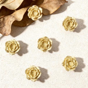 Peut inclure: Six boutons décoratifs dorés en forme de rose sont disposés sur une surface texturée blanc cassé. Les boutons sont détaillés avec de multiples pétales et sont espacés uniformément. Une feuille brune séchée est dans le coin supérieur gauche.