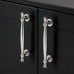 Peut inclure: Gros plan sur deux poignées de placard argentées polies, montées sur une porte de placard noire. Chaque poignée présente un design décoratif en haut et en bas, avec une longue barre incurvée au milieu.