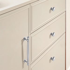 May include: Close-up of a cream-colored cabinet with three drawers and a door. The cabinet features polished chrome hardware, including a long handle and round knobs. The drawers and door have recessed paneling.