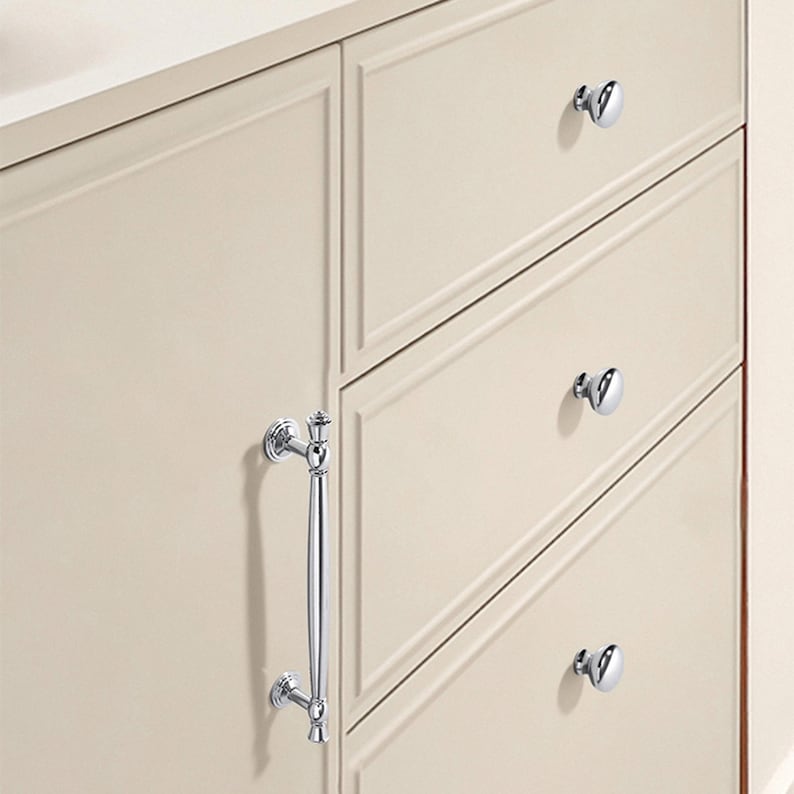 May include: Close-up of a cream-colored cabinet with three drawers and a door. The cabinet features polished chrome hardware, including a long handle and round knobs. The drawers and door have recessed paneling.