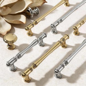Może przedstawiać: Asortyment złotych i srebrnych uchwytów i gałek do szafek. Okucia mają klasyczny design z zaokrąglonymi podstawami i dekoracyjnymi detalami. Złote okucia mają polerowane wykończenie, a srebrne szczotkowane. Ułożone na jasnej powierzchni.