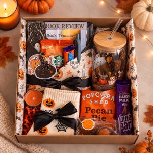 May include: A cardboard gift box containing a glass tumbler with a bamboo lid and straw, cream-coloured socks featuring orange pumpkins and black spiderwebs, a bag of Popcorn Shed Pecan Pie, a Cadbury Dairy Milk bar, a candle, and a printed book tracker.