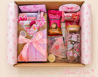 May include: A cardboard gift box lined with pink bow-patterned tissue paper containing a book wrapped in pink striped paper with a large satin bow, a Mallow & Marsh snack, Jammie Dodgers biscuits, a glass tumbler with pink graphics, and a small candle.