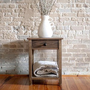 May include: A small, wooden nightstand with a drawer and a lower shelf. A white vase with two handles holds tall, dried branches. A folded blanket rests on the shelf. The background is a light-colored brick wall.