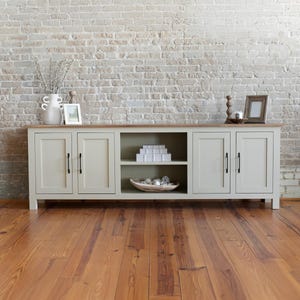 May include: A light green wooden cabinet with four doors and a central open shelf, set against a white brick wall. The cabinet has a light brown top and black handles. The room has a wooden floor.