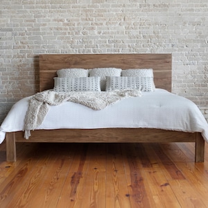 May include: A wooden bed frame with a headboard, covered with a white comforter and pillows. A beige throw blanket is draped over the bed. The bed is set against a white brick wall and a wooden floor.