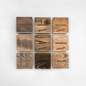 Puede incluir: Nueve posavasos cuadrados de madera con varios patrones de vetas y texto grabado. Los posavasos est&aacute;n etiquetados con tipos de madera como "Smoked Ash" y "Dark Hickory". Cada posavasos tiene el texto "AB Handcrafted" grabado.