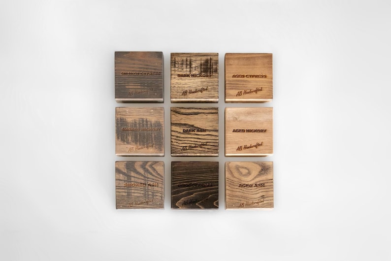 Puede incluir: Nueve posavasos cuadrados de madera con varios patrones de vetas y texto grabado. Los posavasos est&aacute;n etiquetados con tipos de madera como "Smoked Ash" y "Dark Hickory". Cada posavasos tiene el texto "AB Handcrafted" grabado.