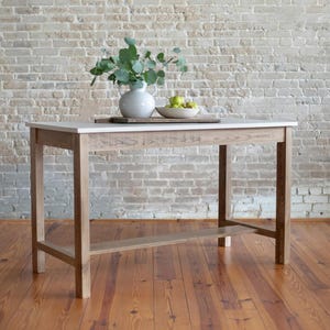 May include: A light-colored wooden kitchen island with a white countertop. The island has a lower shelf and is decorated with a vase of greenery and a bowl of green fruit. The background is a brick wall and a wooden floor.