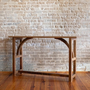 May include: A wooden console table with a natural finish. The table features a rectangular top and arched supports, with a lower horizontal support. The background is a brick wall and a wooden floor.