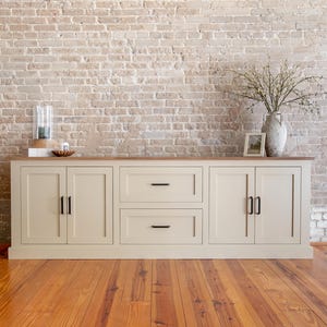 May include: A cream-colored wooden sideboard with black hardware, featuring two sets of doors and two drawers. The sideboard sits on a wooden floor in front of a brick wall. Decorative items are on top.