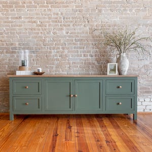 May include: A sage green cabinet with two sets of drawers and two doors, topped with a light wood surface. The cabinet has brass knobs and sits on a wooden floor. A vase with branches, a framed picture, and decorative items are on top.