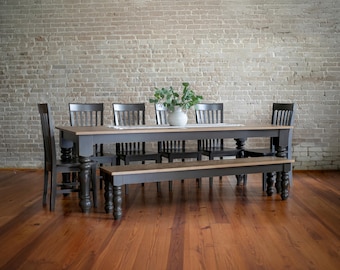 May include: A farmhouse-style dining set with a rectangular table, bench, and chairs. The table and bench have a two-tone design with a dark gray base and a light wood top. The chairs are black. A white vase with greenery sits on the table.