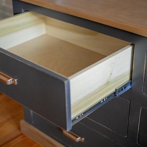 Puede incluir: Una c&oacute;moda gris oscuro con un caj&oacute;n parcialmente abierto. El interior del caj&oacute;n es de madera clara y el exterior tiene tiradores de color cobre. La parte superior es de un tono de madera m&aacute;s claro.