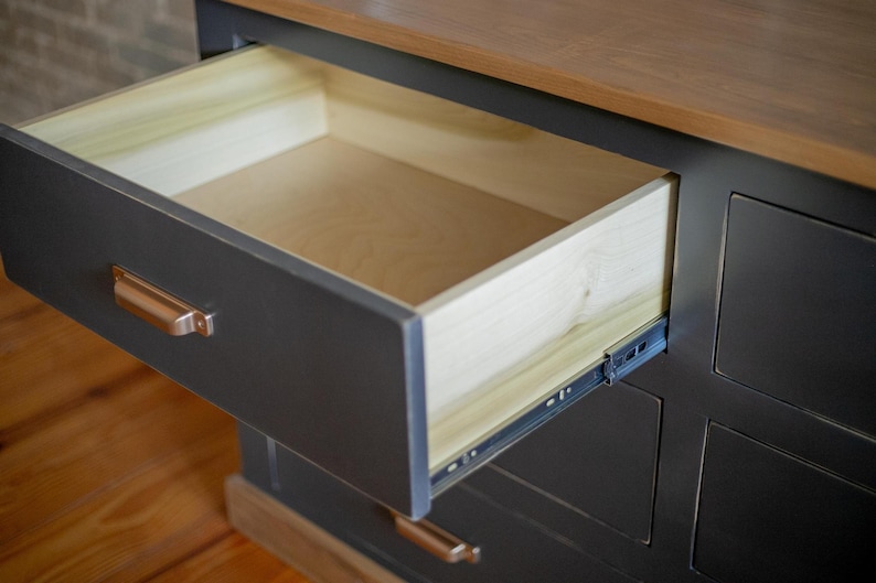Puede incluir: Una c&oacute;moda gris oscuro con un caj&oacute;n parcialmente abierto. El interior del caj&oacute;n es de madera clara y el exterior tiene tiradores de color cobre. La parte superior es de un tono de madera m&aacute;s claro.
