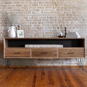 May include: A mid-century modern style wooden media console with three drawers and open shelving. The console has black hairpin legs and is set against a brick wall. Decorative items are displayed on top.
