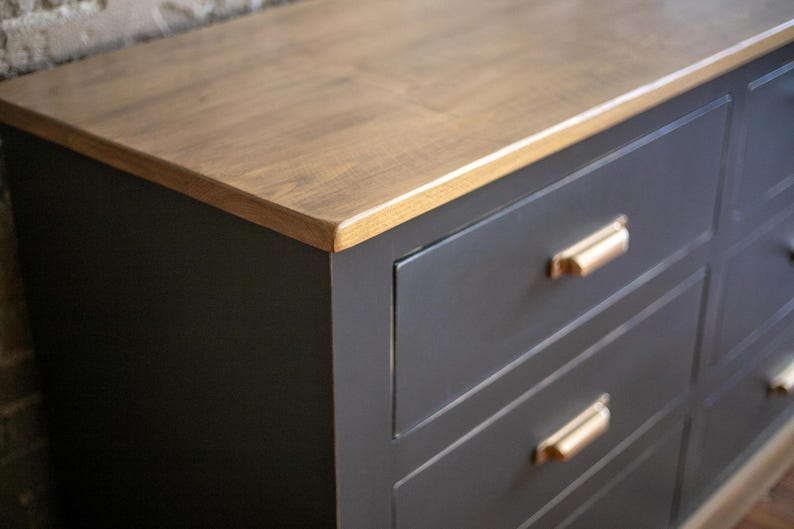 Puede incluir: Una c&oacute;moda gris oscuro con una tapa de madera marr&oacute;n claro. Tiene seis cajones con tiradores dorados. La tapa de madera tiene un patr&oacute;n de vetas natural. La c&oacute;moda es de estilo moderno.