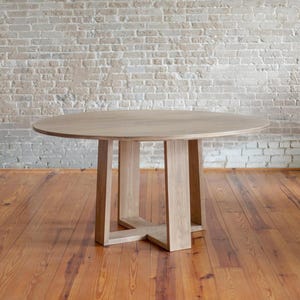 May include: A round wooden table with a light brown finish. The table has a four-legged base with an X-shaped support. The table is set on a wooden floor in front of a brick wall.
