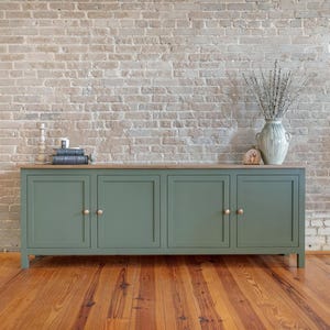 May include: A sage green cabinet with four doors and gold knobs, set against a brick wall and wooden floor. The cabinet has a light wood top. A vase with dried plants and decorative items sit on top.