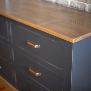 Puede incluir: Una c&oacute;moda gris oscuro con seis cajones y una tapa de madera marr&oacute;n claro. Los cajones tienen tiradores de color cobre. Un mueble moderno.