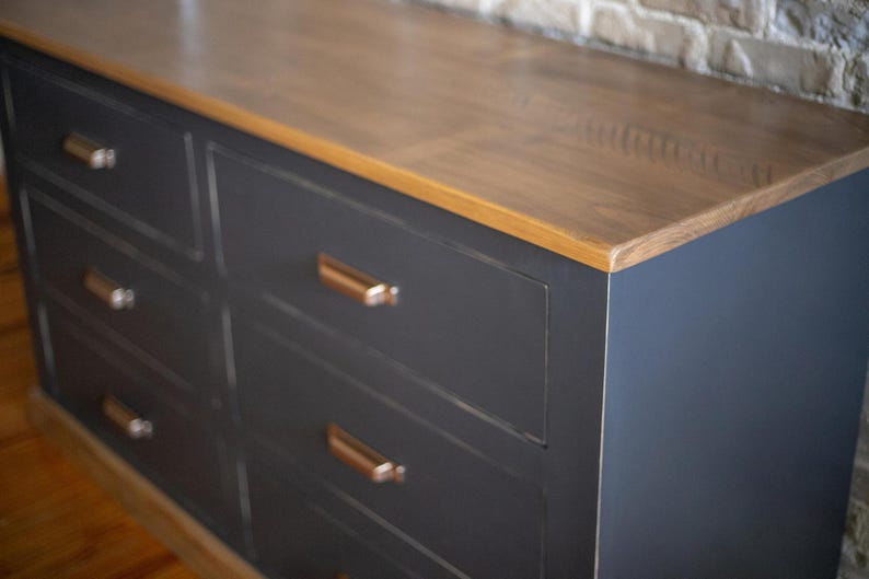 Puede incluir: Una c&oacute;moda gris oscuro con seis cajones y una tapa de madera marr&oacute;n claro. Los cajones tienen tiradores de color cobre. Un mueble moderno.