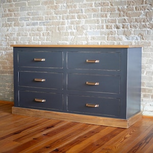 Puede incluir: C&oacute;moda de seis cajones con acabado gris oscuro y detalles en madera clara. Los cajones tienen tiradores color bronce. La parte superior e inferior son de madera clara. La c&oacute;moda est&aacute; colocada contra una pared de ladrillo. Mide unos 152 cm de ancho.