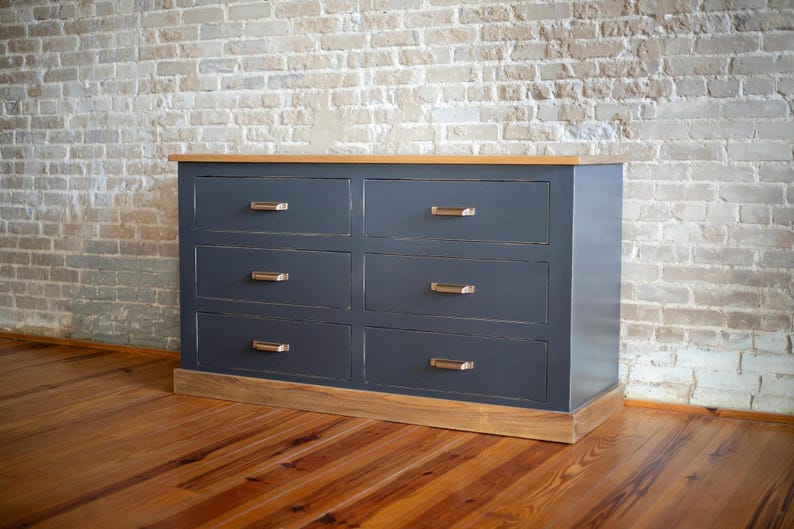 Puede incluir: C&oacute;moda de seis cajones con acabado gris oscuro y detalles en madera clara. Los cajones tienen tiradores color bronce. La parte superior e inferior son de madera clara. La c&oacute;moda est&aacute; colocada contra una pared de ladrillo. Mide unos 152 cm de ancho.