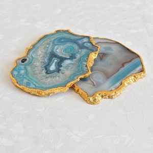 May include: Two agate coasters with gold-painted edges. The coasters display shades of blue, grey, and white, with a natural, organic shape. The gold edges add a touch of elegance to the coasters.