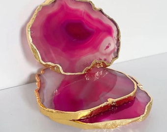 May include: Three pink agate coasters with gold-painted edges. The coasters have a natural, organic shape and a polished surface. The pink color varies in shade, creating a layered effect. These decorative coasters are ideal for protecting surfaces from drink rings.