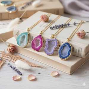 Peut inclure: Quatre colliers avec pendentifs en pierres précieuses et chaînes dorées. Les pendentifs sont dans les tons de bleu, rose et violet, avec des bordures dorées. Les colliers sont exposés sur une pile de livres, avec des fleurs séchées et des pétales éparpillés.