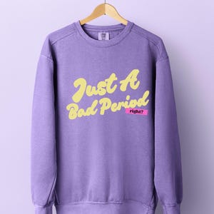 May include: A lavender crewneck sweatshirt featuring the yellow text Just A Bad Period with the word right? printed in white on a small pink rectangular background. The garment has long sleeves, a ribbed neckline, and ribbed cuffs and hem.
