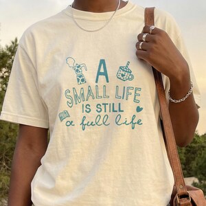 Könnte beinhalten: Cremefarbenes T-Shirt mit dem Text "A small life is still a full life" in Türkis. Das Shirt zeigt kleine Illustrationen einer Kaffeetasse, eines Buches und anderer Objekte. Die Person trägt eine hellblaue Jeans und braune Sandalen.