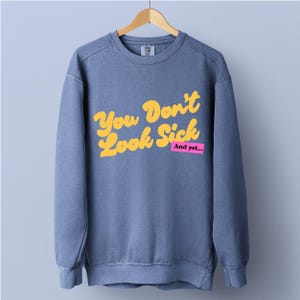 Könnte beinhalten: Ein blaues Sweatshirt mit Rundhalsausschnitt und dem gelben Schriftzug "You Don't Look Sick" in einer Retro-Schriftart, ergänzt durch den Text "And yet..." in Weiß auf einem kleinen rosa Rechteck unter dem Hauptsatz.