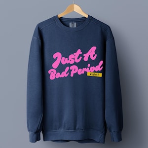 May include: A navy blue crewneck sweatshirt featuring the pink text Just A Bad Period in a cursive font, with the word right? printed in black on a yellow rectangular background located below the main text on the front of the garment.