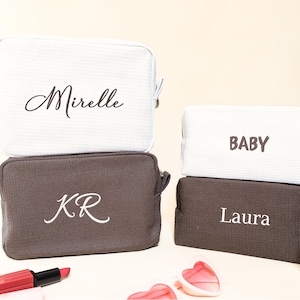 May include: Four personalized cosmetic bags in white and brown. The white bags have the names "Mirelle" and "BABY" embroidered on them. The brown bags have the initials "KR" and the name "Laura" embroidered on them. Each bag has a zipper closure.