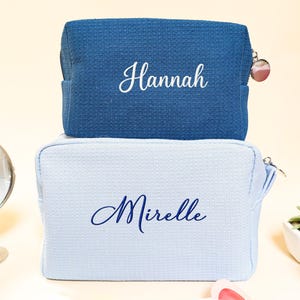 May include: Two personalized cosmetic bags. The top bag is a textured navy blue, with the name "Hannah" embroidered in white. The bottom bag is light blue with "Mirelle" embroidered in navy. Both have zipper closures.
